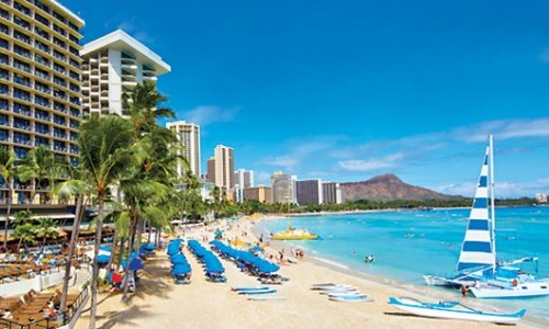 Sunny beach with vacationers, sailboats, and a view of high-rise hotels against a backdrop of a mountain, perfect for Hawaii cruise shore excursions.