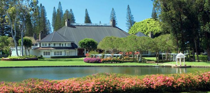 lanai-Koele-lodge a well-manicured estate with a large pond and flowering bushes in the foreground, and a grand residence surrounded by trees in the background.