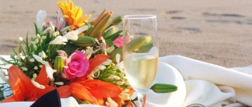 An arrangement of flowers next to a glass of white wine on a beach setting.