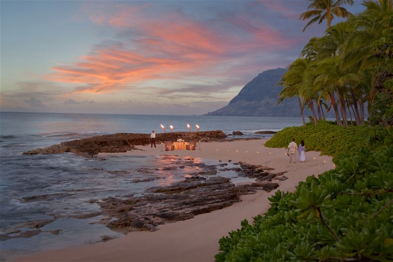 A couple walking along a beach at sunset with a torch-lit dinner setup by the shore.