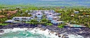 Aerial view of a coastal resort on the Big Island with white buildings and swimming pools, overlooking a rugged shoreline with lush greenery in the background.