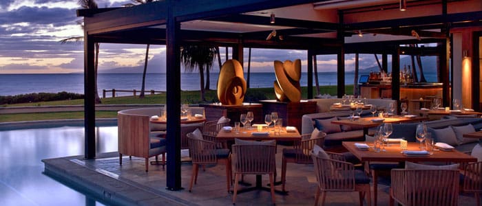 Oceanfront restaurant at dusk with an elegant dining setup and contemporary art sculptures.