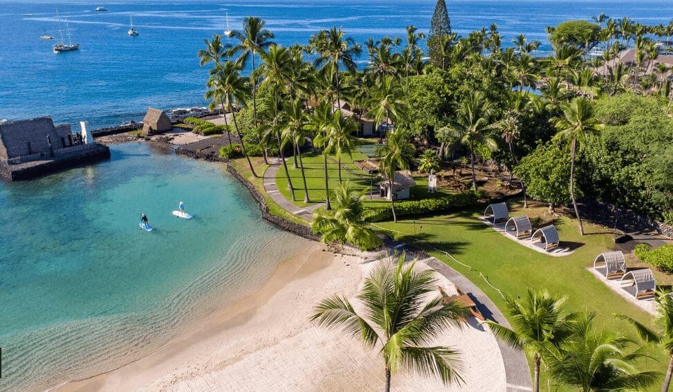 Aerial view of a tranquil tropical beach in Hawaii with palm trees, sun loungers, and people paddleboarding in clear blue waters.